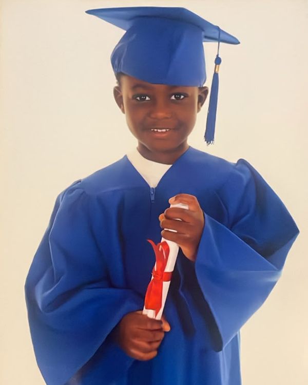Smiling young child in a blue graduation cap and gown holding a rolled certificate with a red ribbon