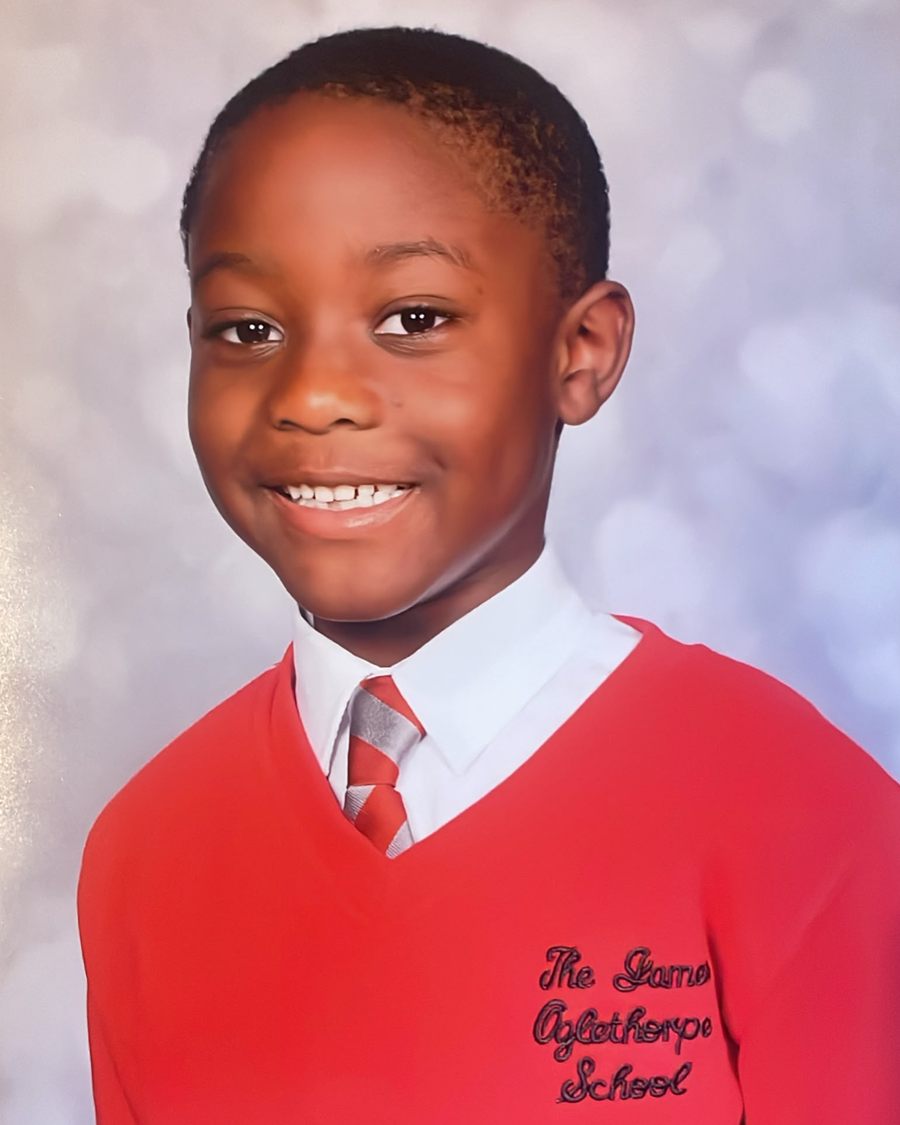 Smiling schoolboy in a red school jumper and striped tie, photographed against a soft grey backdrop